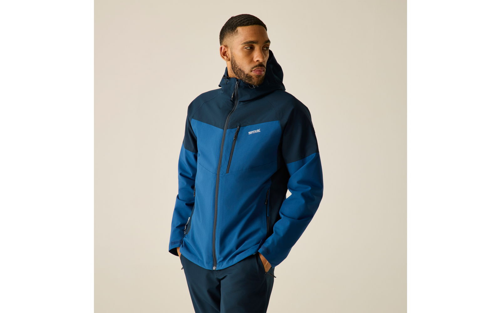 Person in blauer Softshell-Jacke mit Kapuze, steht mit Händen in den Taschen und blickt zur Seite vor hellem, einfarbigem Hintergrund