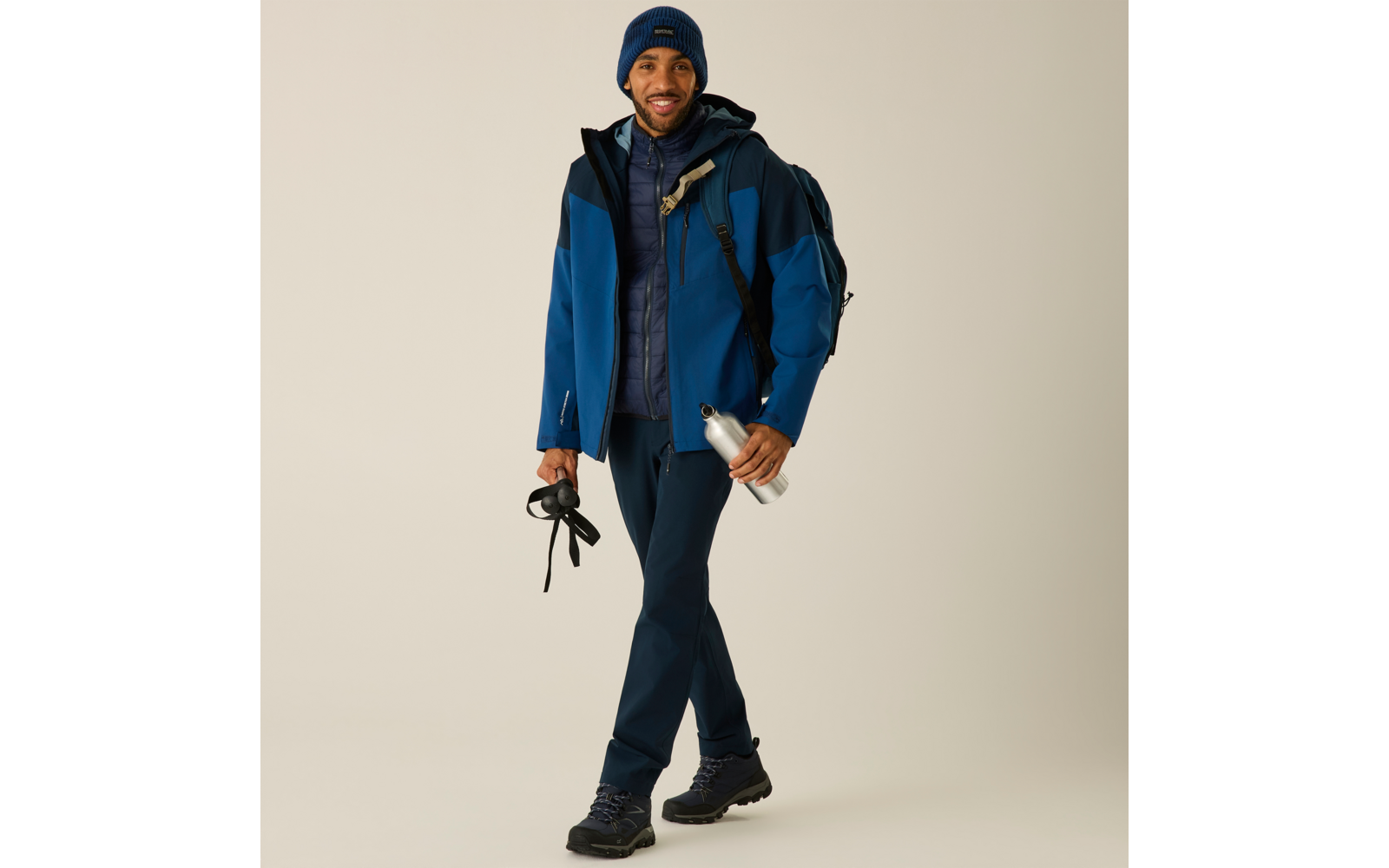 Person in blauer Jacke und Mütze, mit Rucksack, hält Stöcke und eine Metall-Trinkflasche, dunkle Hose und Wanderstiefel, heller einfarbiger Hintergrund