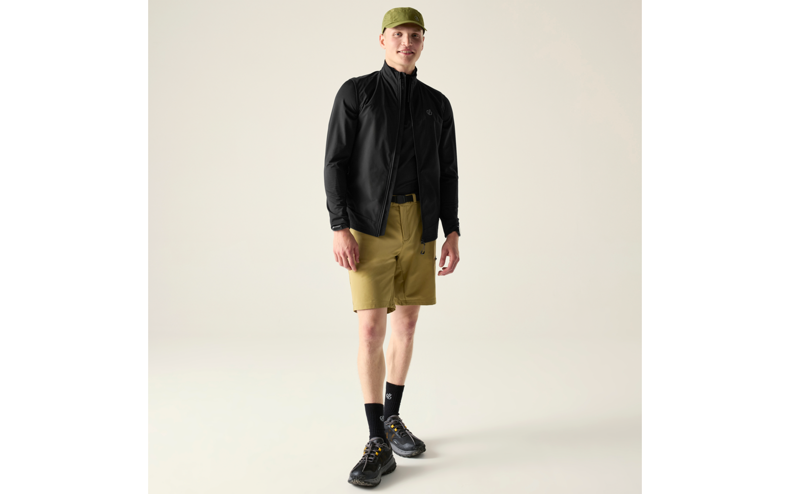 Mann steht, trägt grüne Kappe, schwarze Jacke, khakifarbene Shorts, schwarze Socken und schwarze Sneaker, heller Hintergrund