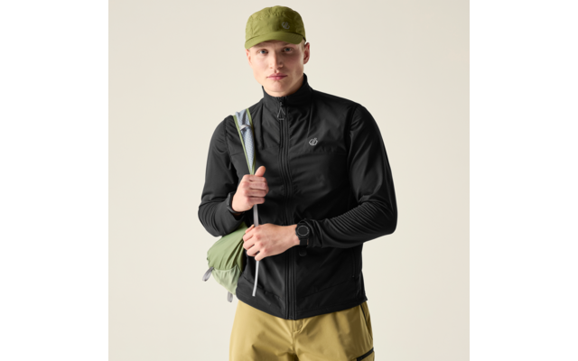 Person frontal, trägt olivgrüne Kappe, schwarze Reißverschlussjacke, hellbraune Shorts, grüner Rucksack über der rechten Schulter, hält den Riemen mit beiden Händen, schwarze Armbanduhr am linken Handgelenk, heller einfarbiger Hintergrund