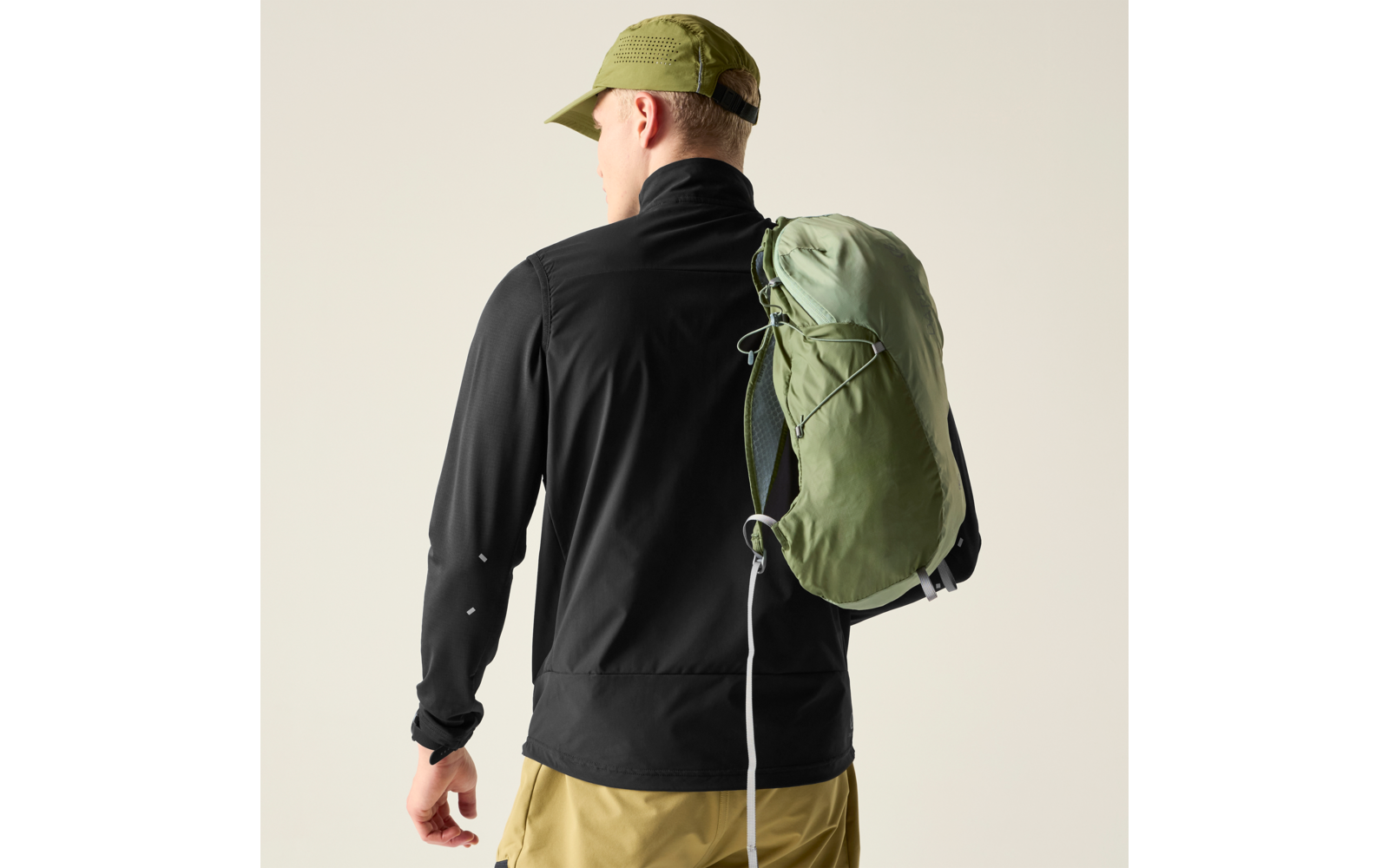 Person von hinten, trägt schwarze Jacke, hellgrünes Cap, khakifarbene Hose und einen grünen Rucksack über einer Schulter