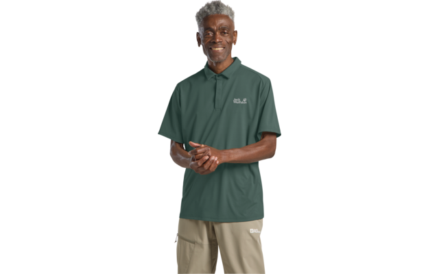 Ein stehender Mann mit kurzen grauen Haaren, trägt ein grünes Poloshirt mit Logo und beige Hose, Hände vor dem Körper gefaltet, schaut in die Kamera