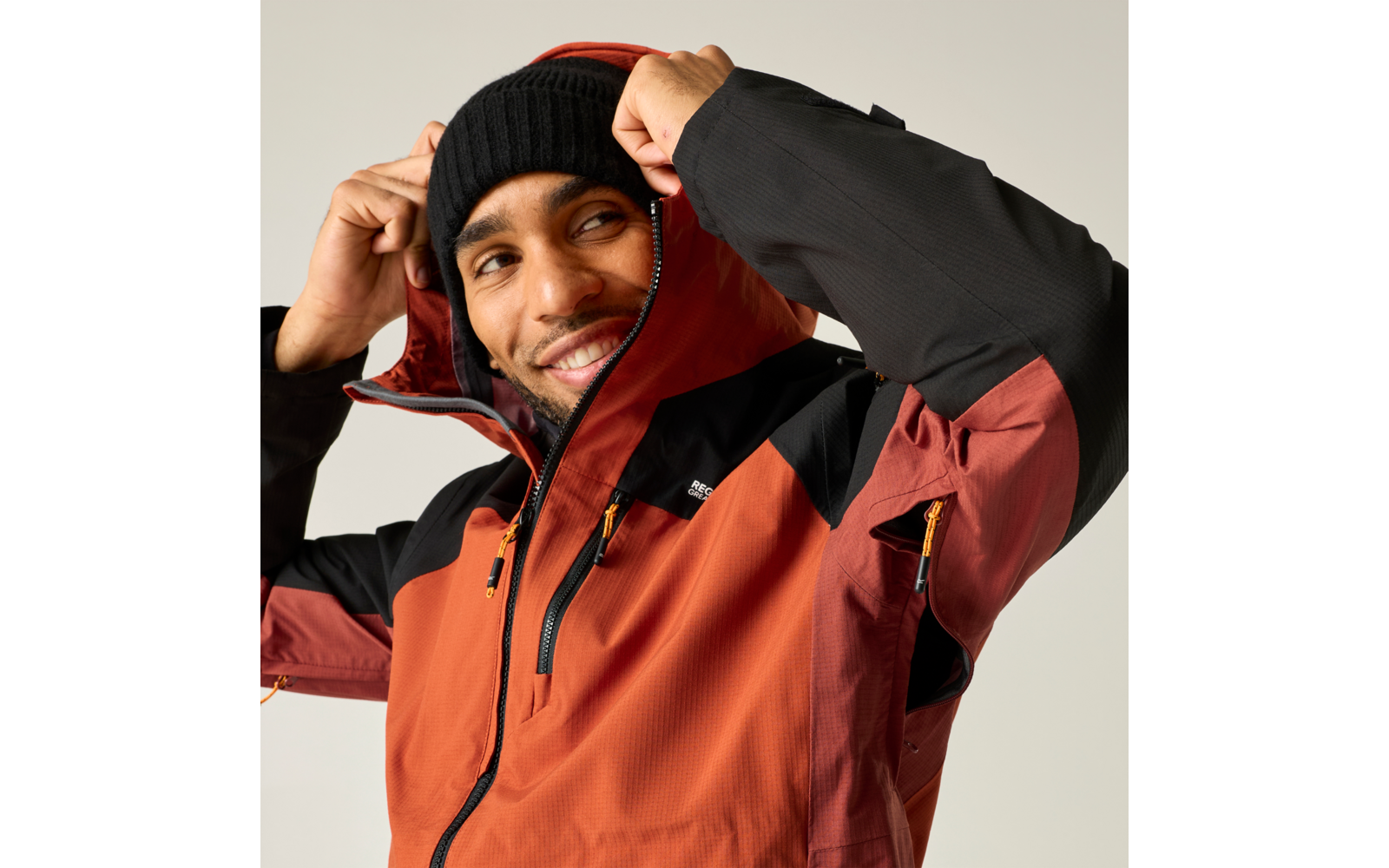 Person zieht Kapuze eines orange-schwarzen Outdoor-Jackes hoch, trägt schwarze Strickmütze und lächelt