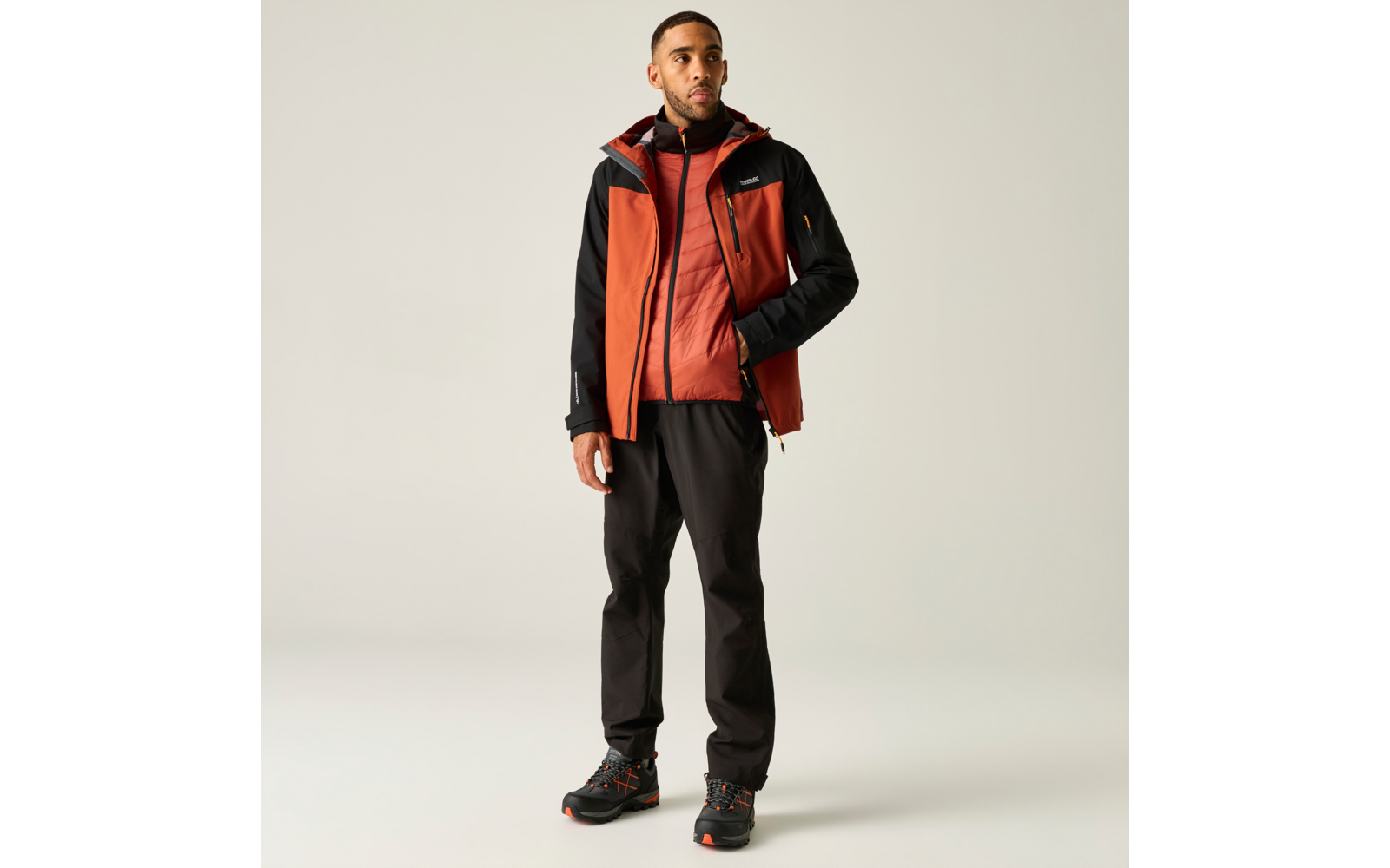 Eine stehende Person in orange-roter Outdoorjacke mit schwarzen Ärmeln, schwarzer Hose und schwarzen Wanderschuhen mit orangefarbenen Details vor hellem Hintergrund