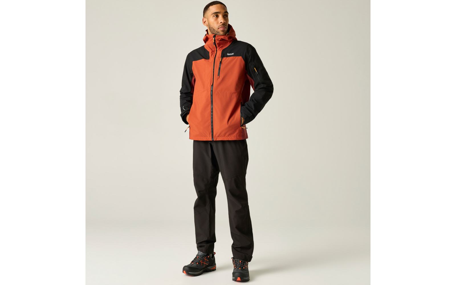 Mann steht, schaut nach rechts, trägt orange-schwarze Kapuzen-Outdoorjacke, schwarze Hose und Wanderschuhe, Hände in den Taschen
