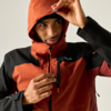 Person in orange-schwarzer Outdoorjacke mit Kapuze zieht am Reißverschluss der Brusttasche, blickt nach rechts