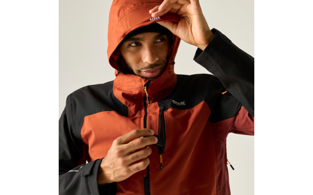 Person in orange-schwarzer Outdoorjacke mit Kapuze zieht am Reißverschluss der Brusttasche, blickt nach rechts