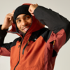 Person zieht Kapuze eines orange-schwarzen Outdoor-Jackes hoch, trägt schwarze Strickmütze und lächelt
