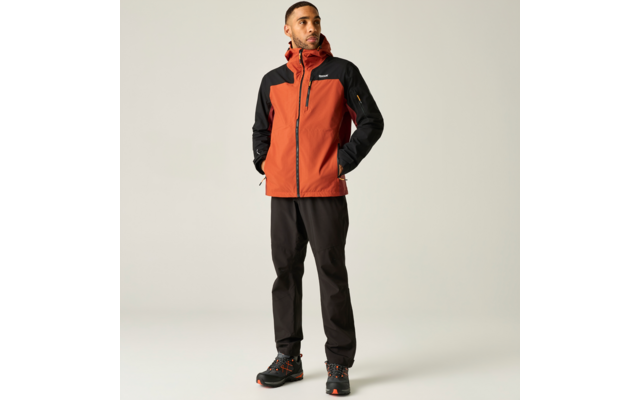 Mann steht, schaut nach rechts, trägt orange-schwarze Kapuzen-Outdoorjacke, schwarze Hose und Wanderschuhe, Hände in den Taschen