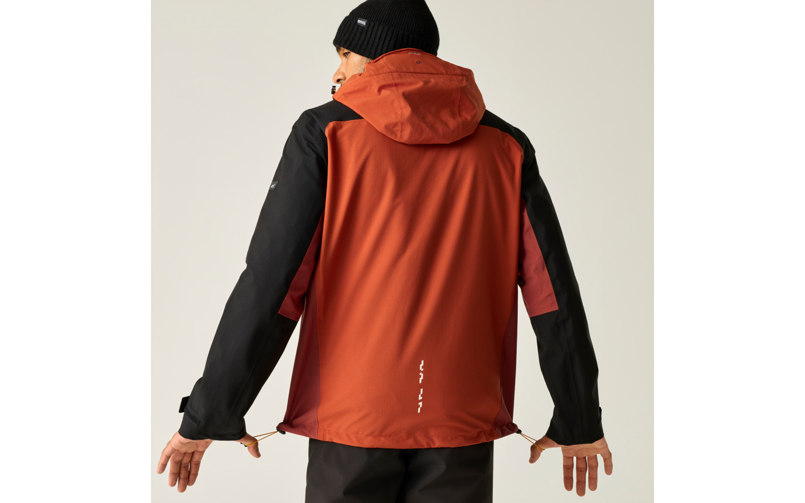Person von hinten in orange-roter Jacke mit schwarzen Ärmeln und Kapuze und schwarzer Mütze, zieht an den Kordeln am Saum, vor hellem Hintergrund