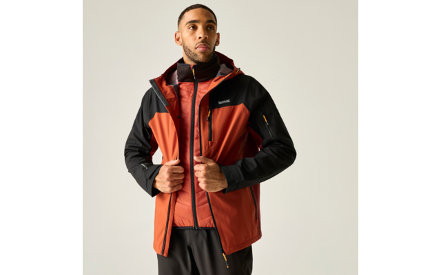 Mann vor hellem Hintergrund, trägt orange-schwarze Outdoorjacke über roter gesteppter Weste, hält Vorderseite der Jacke mit beiden Händen