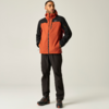 Mann steht, schaut nach rechts, trägt orange-schwarze Kapuzen-Outdoorjacke, schwarze Hose und Wanderschuhe, Hände in den Taschen
