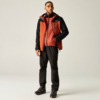 Eine stehende Person in orange-roter Outdoorjacke mit schwarzen Ärmeln, schwarzer Hose und schwarzen Wanderschuhen mit orangefarbenen Details vor hellem Hintergrund