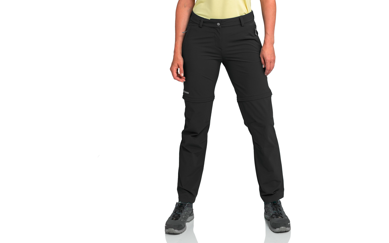 Unterkörper einer Person mit schwarzer Hose mit Reißverschlüssen an den Knien, grauen Wanderschuhen, sichtbarem gelben Oberteilansatz, Händen an den Seiten, weißem Hintergrund