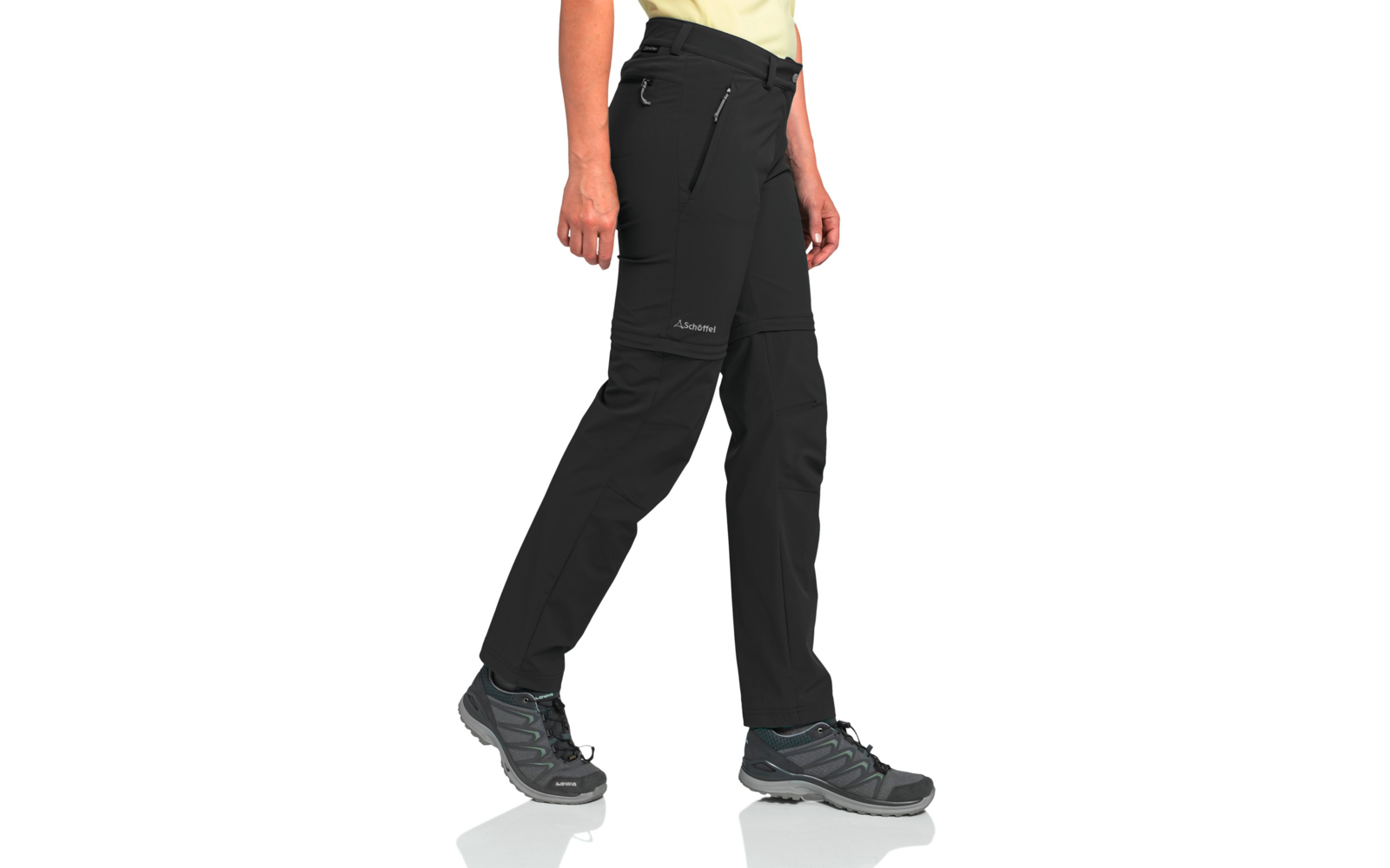 Unterkörper einer stehenden Person mit schwarzer Outdoorhose mit Reißverschlusstaschen, grauen Wanderschuhen und sichtbarem Saum eines gelben Oberteils, Hände seitlich