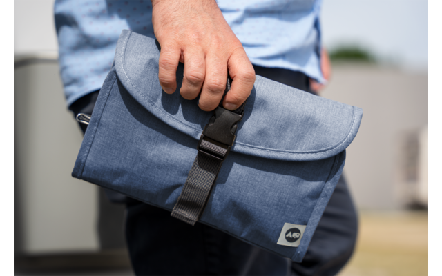 A person's hand holding a blue fabric pouch with a black buckle; the person wears a light blue shirt and dark pants; blurred background
