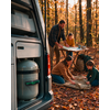 Open camper met zichtbare gasfles; twee volwassenen en twee kinderen in een herfstbos, één wijst op een kaart op een klaptafel, twee leggen een slaapzak uit; rugzak en kampeerservies zichtbaar