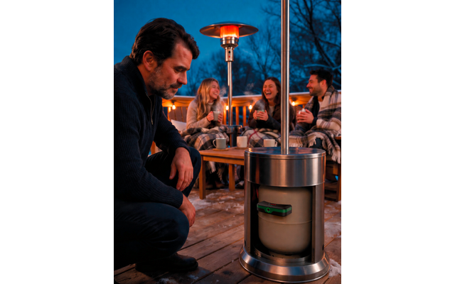 Een man knielt naast een metalen terrasverwarmer met de gasfles zichtbaar; op de achtergrond zitten drie personen op een houten terras, in dekens gewikkeld en met mokken in hun handen, lichtslingers en kale bomen tegen een donkere lucht