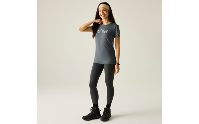 Stehende Person vor hellem Hintergrund, trägt beige Stirnband, graues T‑Shirt mit weißem Aufdruck, schwarze Leggings, schwarze Schnürstiefel und Armbanduhr; lächelnd, rechte Hand am Hals