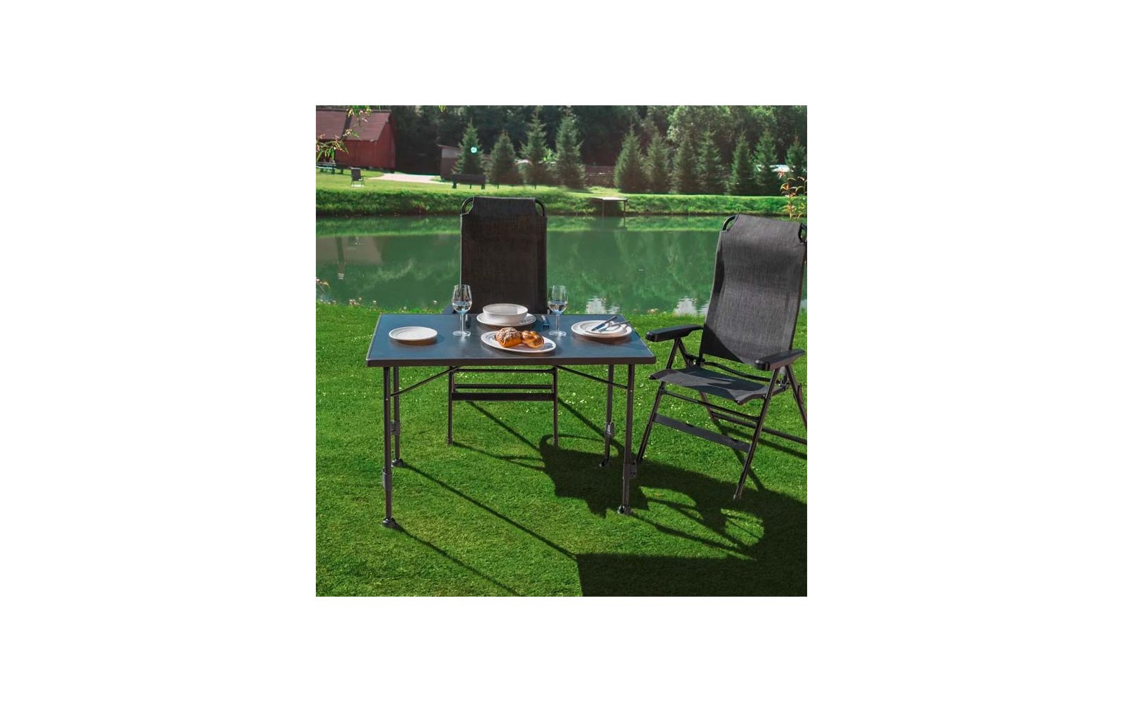 Zwei schwarze Klappstühle und ein Tisch auf Rasen am Teich; Teller, Gläser, Gebäck, Besteck, Bäume und rotes Haus im Hintergrund