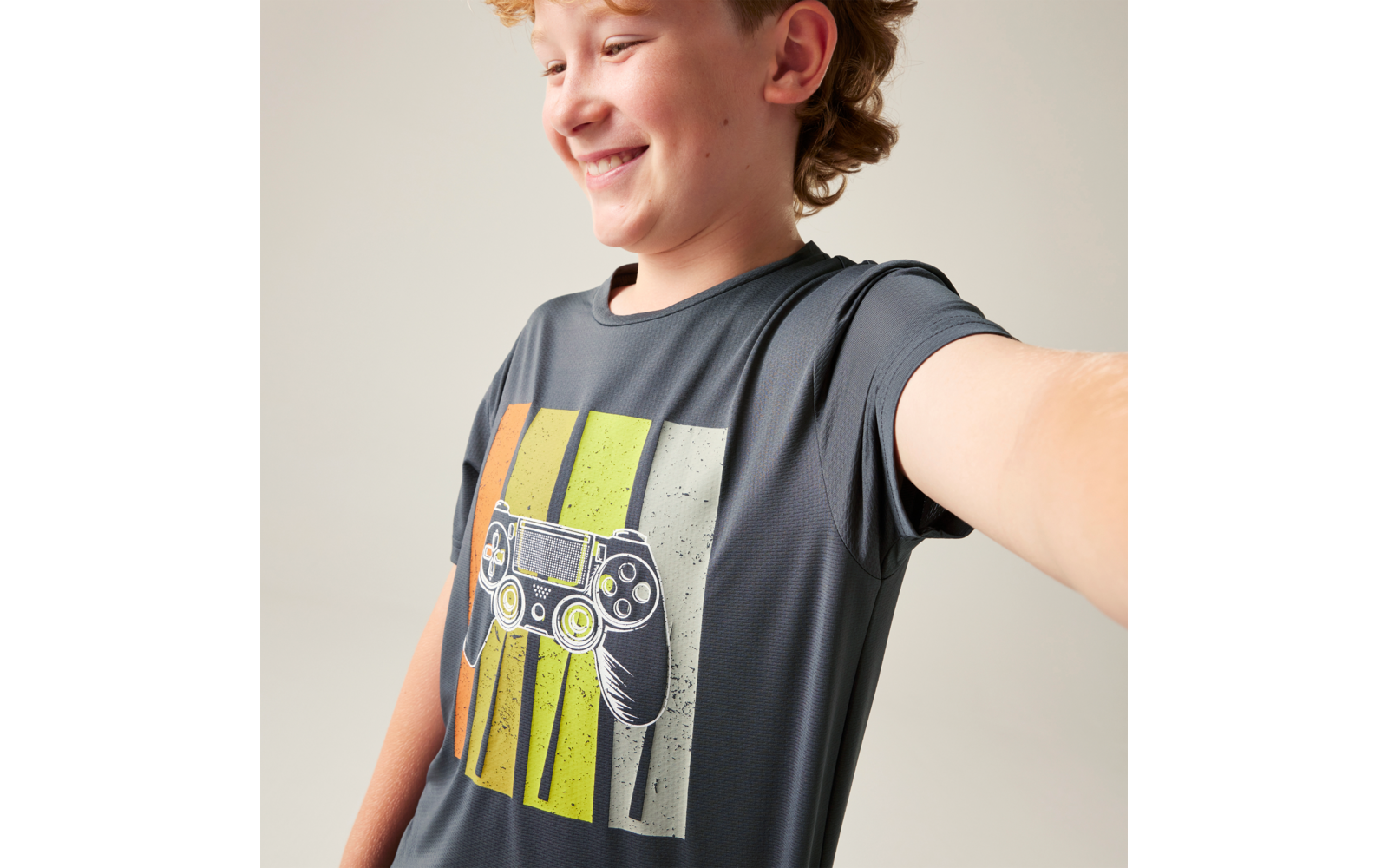 Lächelnder Junge, halb im Bild, ausgestreckter Arm, dunkles T-Shirt mit aufgedrucktem Videospiel-Controller und senkrechten orange-gelben Streifen