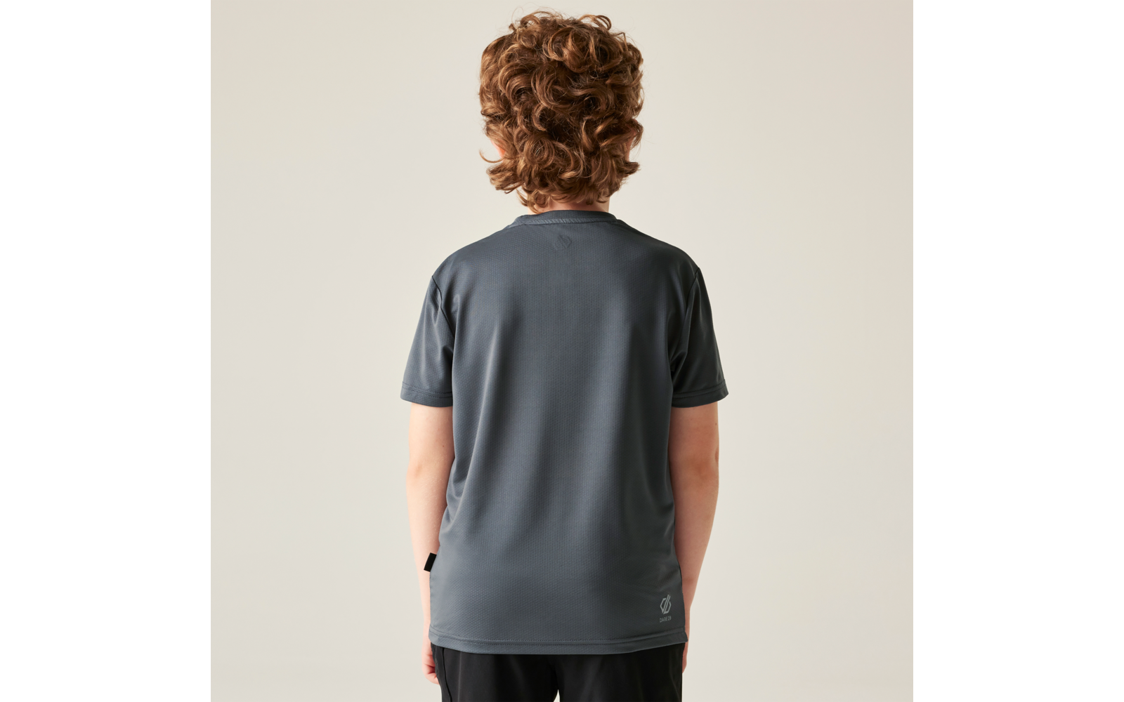 Person von hinten mit schulterlangen lockigen Haaren, grauem T-Shirt und schwarzen Shorts vor hellem Hintergrund