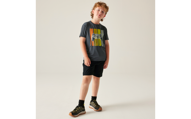 Junge mit lockigen roten Haaren steht vor hellem Hintergrund, trägt dunkles T‑Shirt mit bunten vertikalen Streifen und weißem Aufdruck, schwarze Shorts und grün‑braune Sneaker