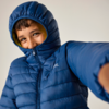 Person in blauer gesteppter Jacke mit hochgezogener Kapuze, lächelnd, Blick zur Kamera, ausgestreckter Arm im Vordergrund