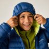 Kind in blauer Steppjacke mit hochgezogener Kapuze, hält die Kapuze mit beiden Händen, trägt grünen Fleecepullover, lächelt