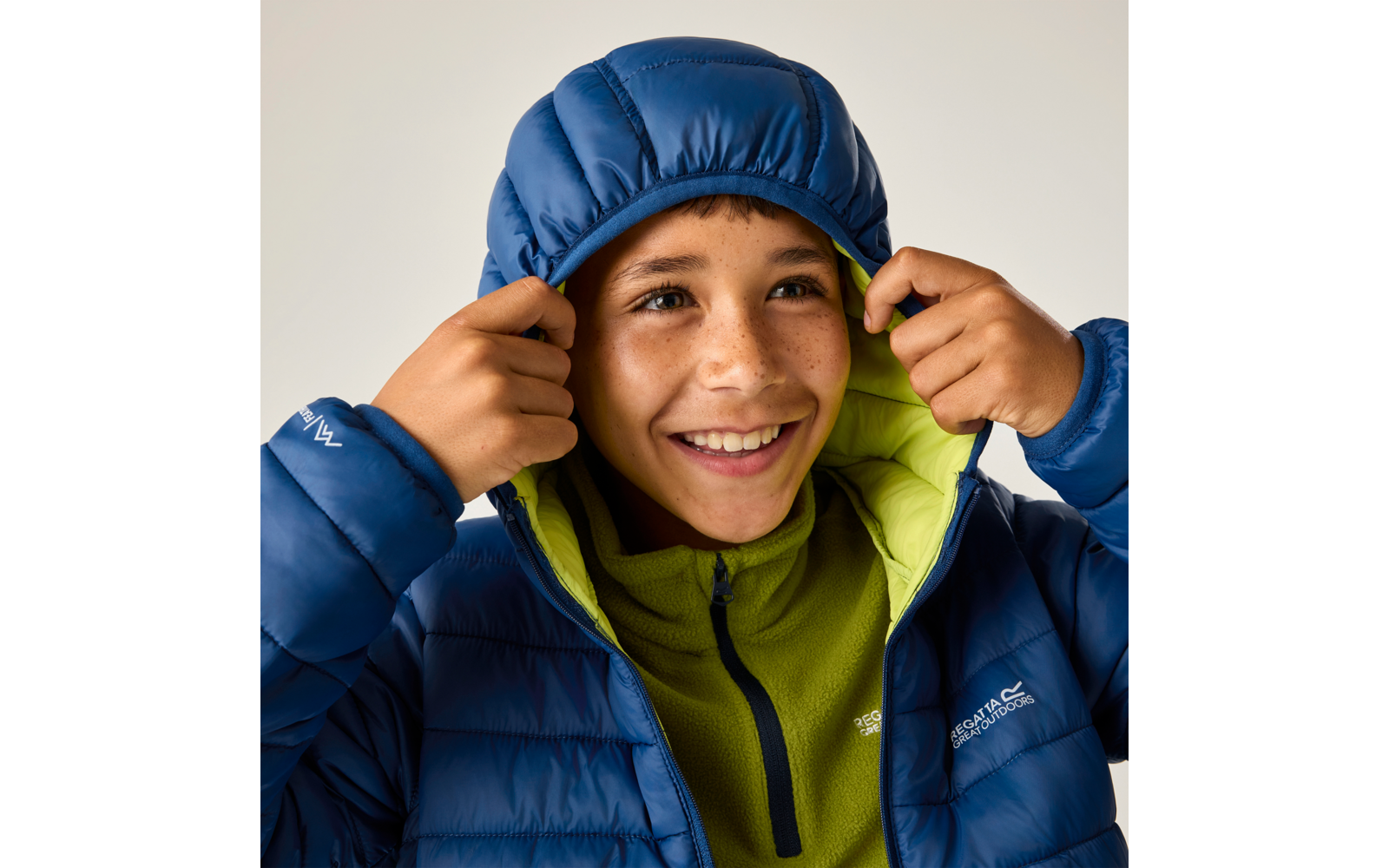 Kind in blauer Steppjacke mit hochgezogener Kapuze, hält die Kapuze mit beiden Händen, trägt grünen Fleecepullover, lächelt