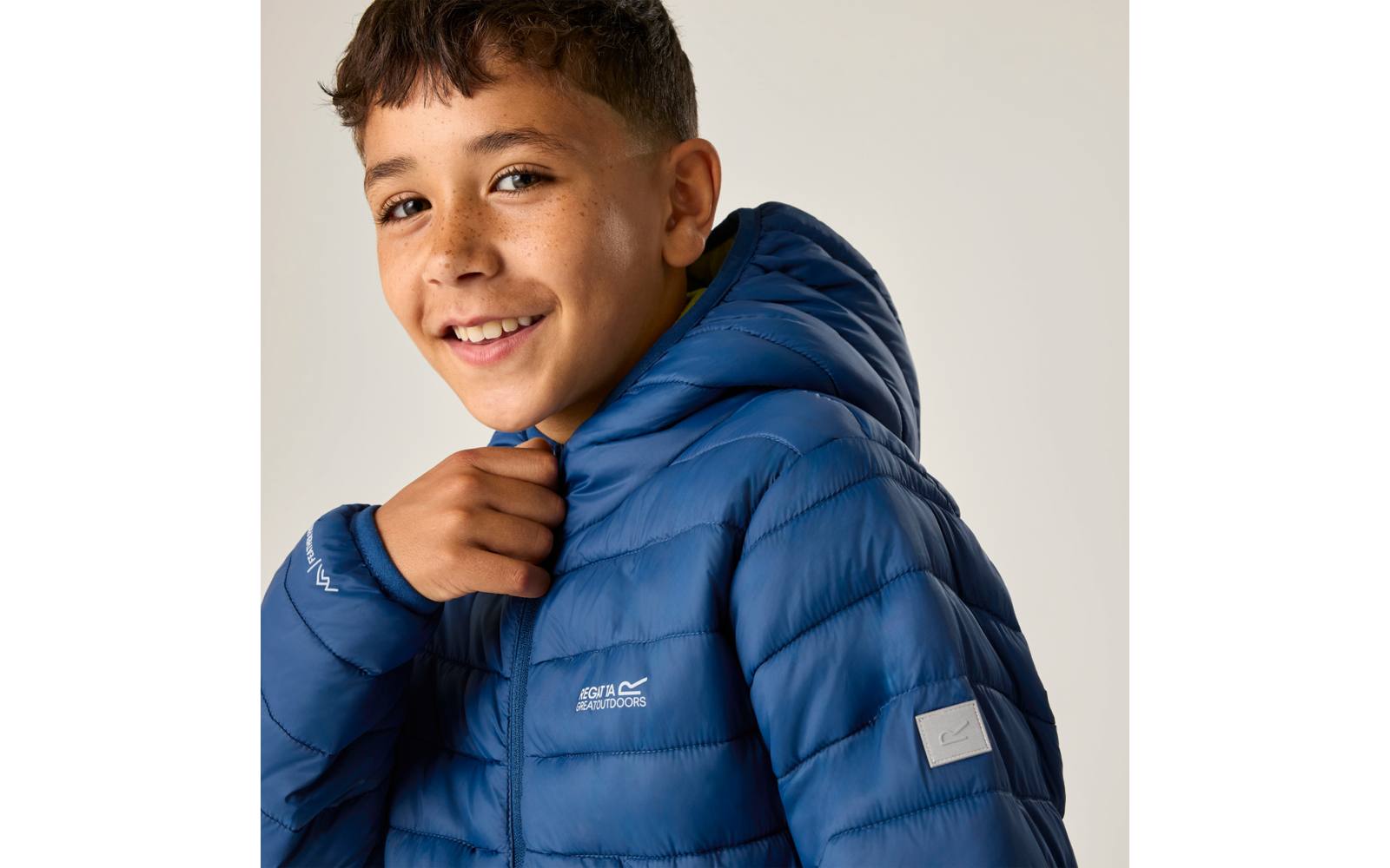 Nahaufnahme eines lächelnden Jungen mit blauer Steppjacke und Kapuze, er hält den Kragen, heller Hintergrund