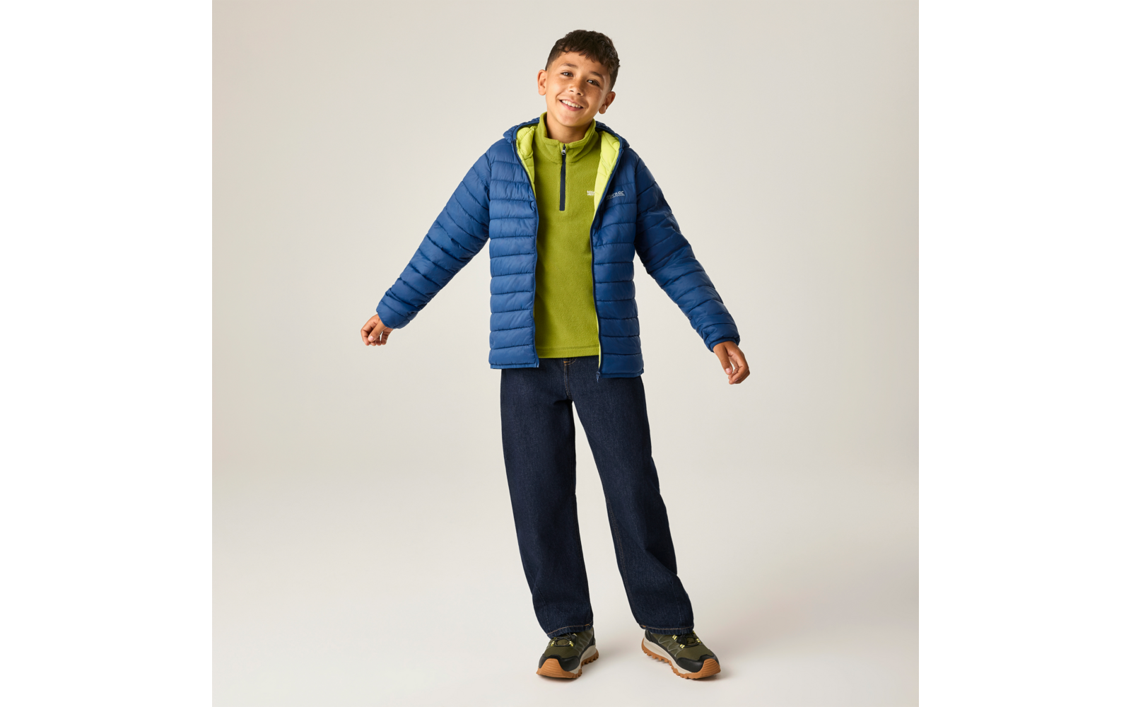 Lächelnder Junge steht mit leicht gespreizten Armen, trägt blaue Steppjacke, grüne Kapuzenjacke, dunkle Jeans und Sneakers vor neutralem Hintergrund