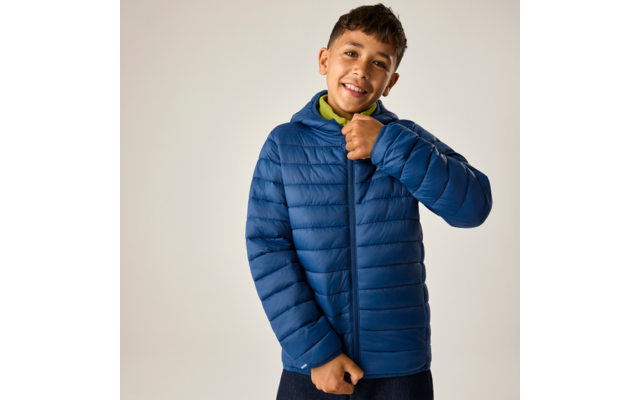Junge mit kurzem dunklem Haar trägt eine blaue gesteppte Jacke und zieht den Reißverschluss hoch; grüner Kapuzenpullover darunter, heller einfarbiger Hintergrund