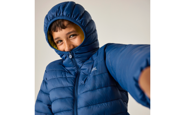 Person in blauer gesteppter Jacke mit hochgezogener Kapuze, lächelnd, Blick zur Kamera, ausgestreckter Arm im Vordergrund
