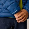 Nahaufnahme ineinandergreifender Hände vor dem Ärmel einer blauen Steppjacke mit dem Schriftzug 'FEATHER FREE', grüner Innenlage und dunkler Jeans