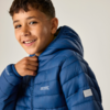 Nahaufnahme eines lächelnden Jungen mit blauer Steppjacke und Kapuze, er hält den Kragen, heller Hintergrund
