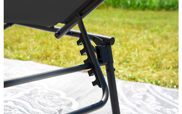 Primo piano del telaio metallico di una sdraio regolabile con tessuto nero, tacche di regolazione, una coperta chiara con motivo sotto e prato sullo sfondo