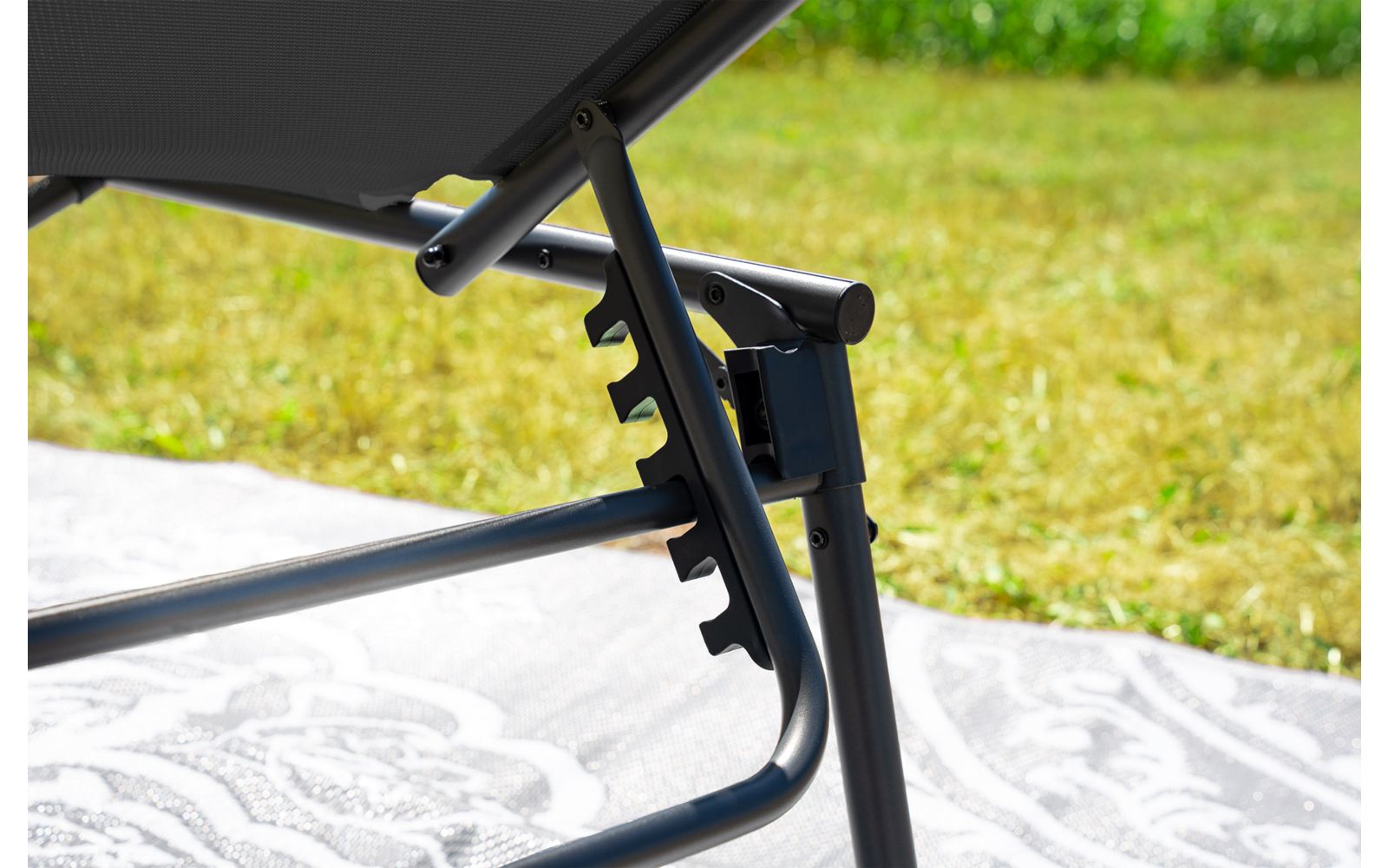 Primo piano del telaio metallico di una sdraio regolabile con tessuto nero, tacche di regolazione, una coperta chiara con motivo sotto e prato sullo sfondo