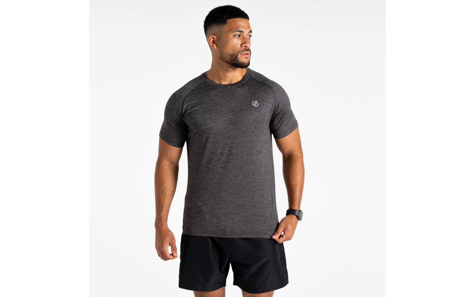Homme debout devant un fond blanc, portant un t‑shirt gris foncé à manches courtes avec petit logo sur la poitrine, un short noir et une montre connectée au poignet gauche, regardant sur le côté