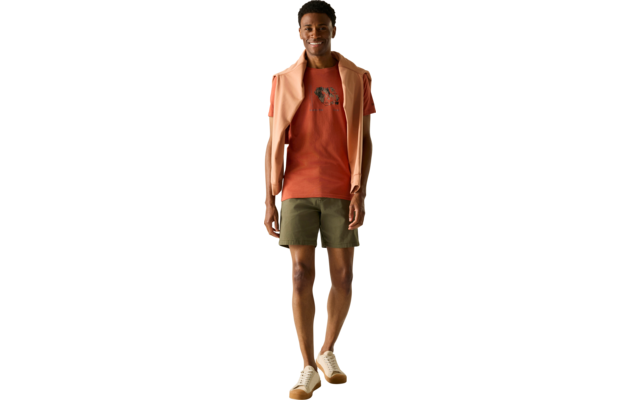 Person geht zur Kamera, trägt oranges T‑Shirt mit Motiv, leichte Jacke über den Schultern, olivgrüne Shorts und helle Sneakers