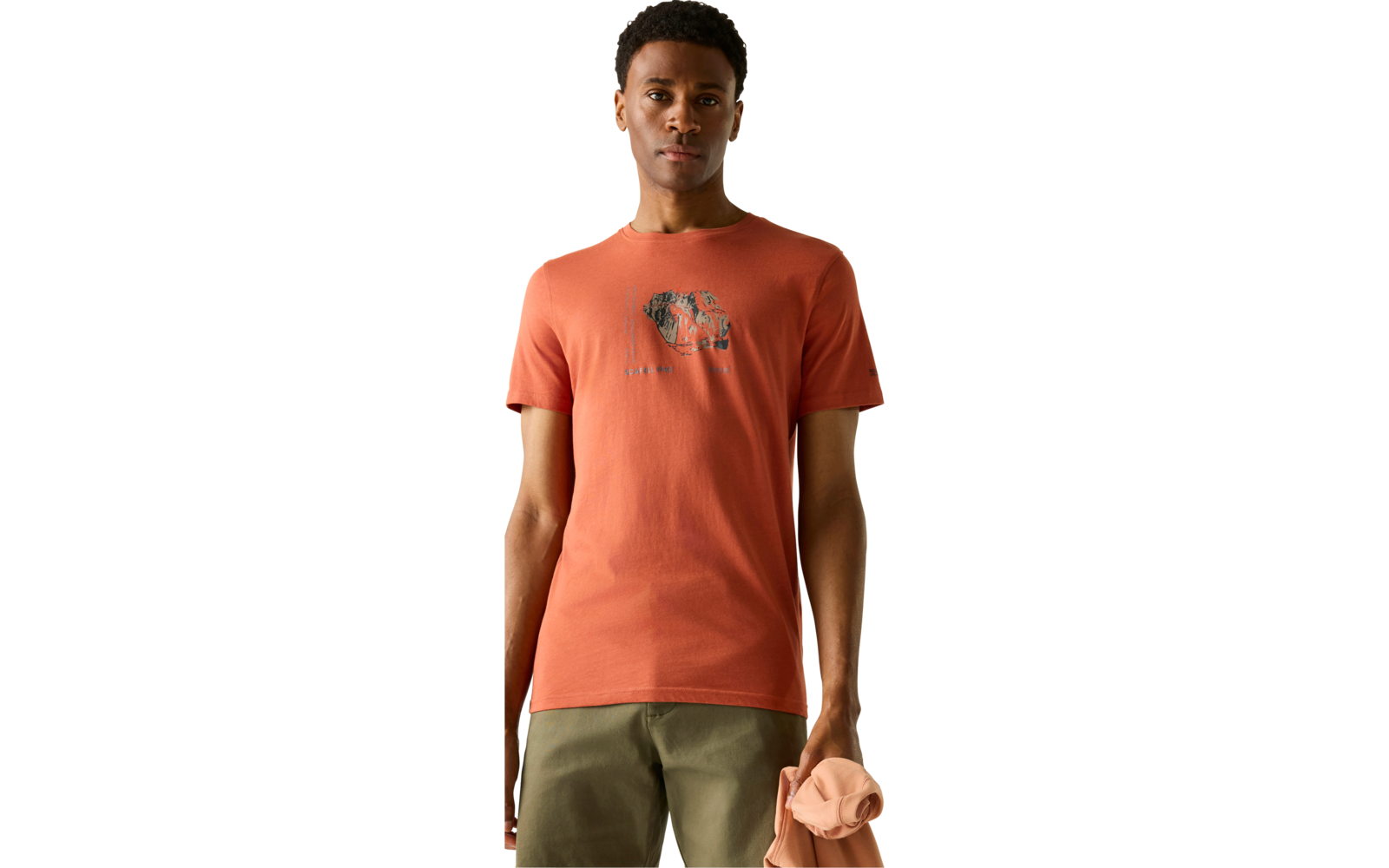 Mann mit kurzem dunklem Haar, orangefarbenes T‑Shirt mit Elefantenprint, olivgrüne Hose, hält einen hellen, gefalteten Pullover; weißer Hintergrund