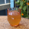 Ein transparentes Glas mit orangefarbenem Getränk, auf einem Mosaiktisch, im Hintergrund grüne Pflanzen