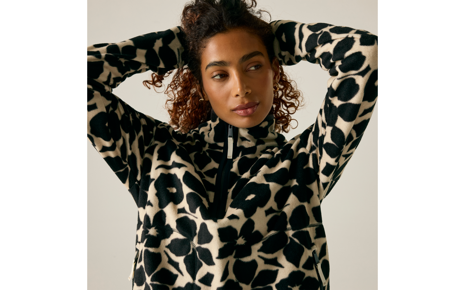 Personne aux cheveux bouclés attachés et boucles d'oreilles dorées, porte un pull motif noir et blanc avec une petite fermeture éclair ; mains derrière la tête, regard de côté, fond clair