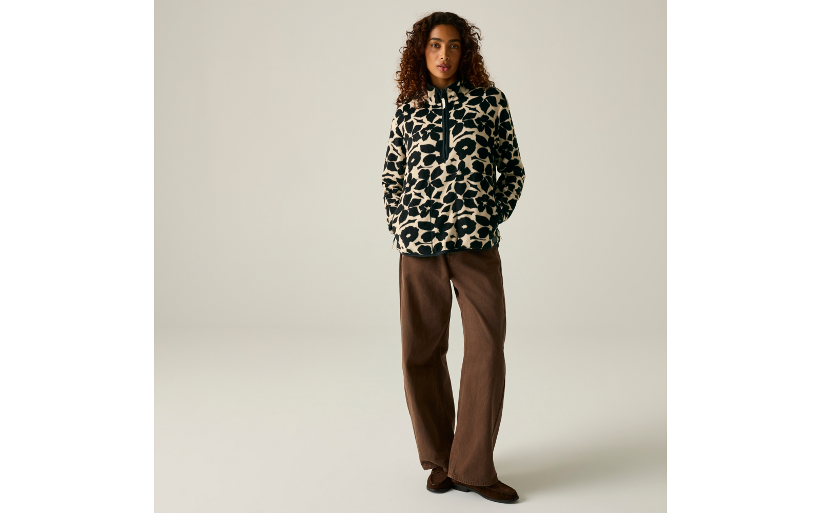 Personne aux cheveux bouclés porte une veste polaire crème à motif de taches noires, mains dans les poches, pantalon brun ample, debout devant un fond uni clair