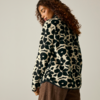 Personne aux cheveux bouclés, vue de dos et légèrement de profil, porte un pull noir et blanc à grand motif et un pantalon marron, fond beige clair