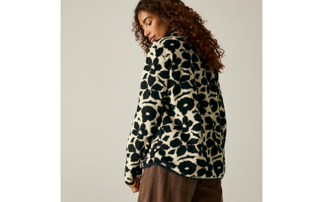 Personne aux cheveux bouclés, vue de dos et légèrement de profil, porte un pull noir et blanc à grand motif et un pantalon marron, fond beige clair