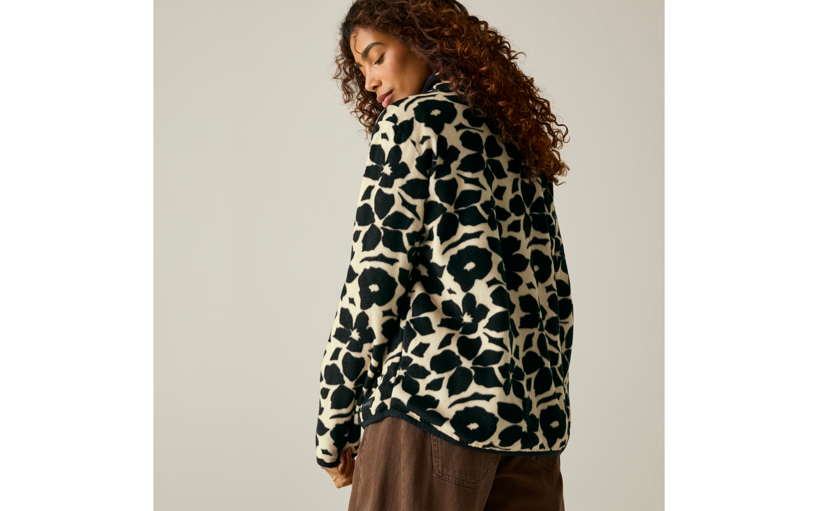 Personne aux cheveux bouclés, vue de dos et légèrement de profil, porte un pull noir et blanc à grand motif et un pantalon marron, fond beige clair