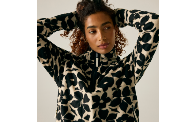 Personne aux cheveux bouclés attachés et boucles d'oreilles dorées, porte un pull motif noir et blanc avec une petite fermeture éclair ; mains derrière la tête, regard de côté, fond clair