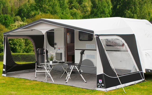 Wohnwagen mit angebautem grauem Vorzelt auf einer Wiese vor bewaldeten Hügeln