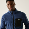 Junger Mann mit blauem Pullover und schwarzer Brusttasche vor hellem Hintergrund